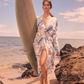 woman poses on beach in linen dress holding a surfboard
